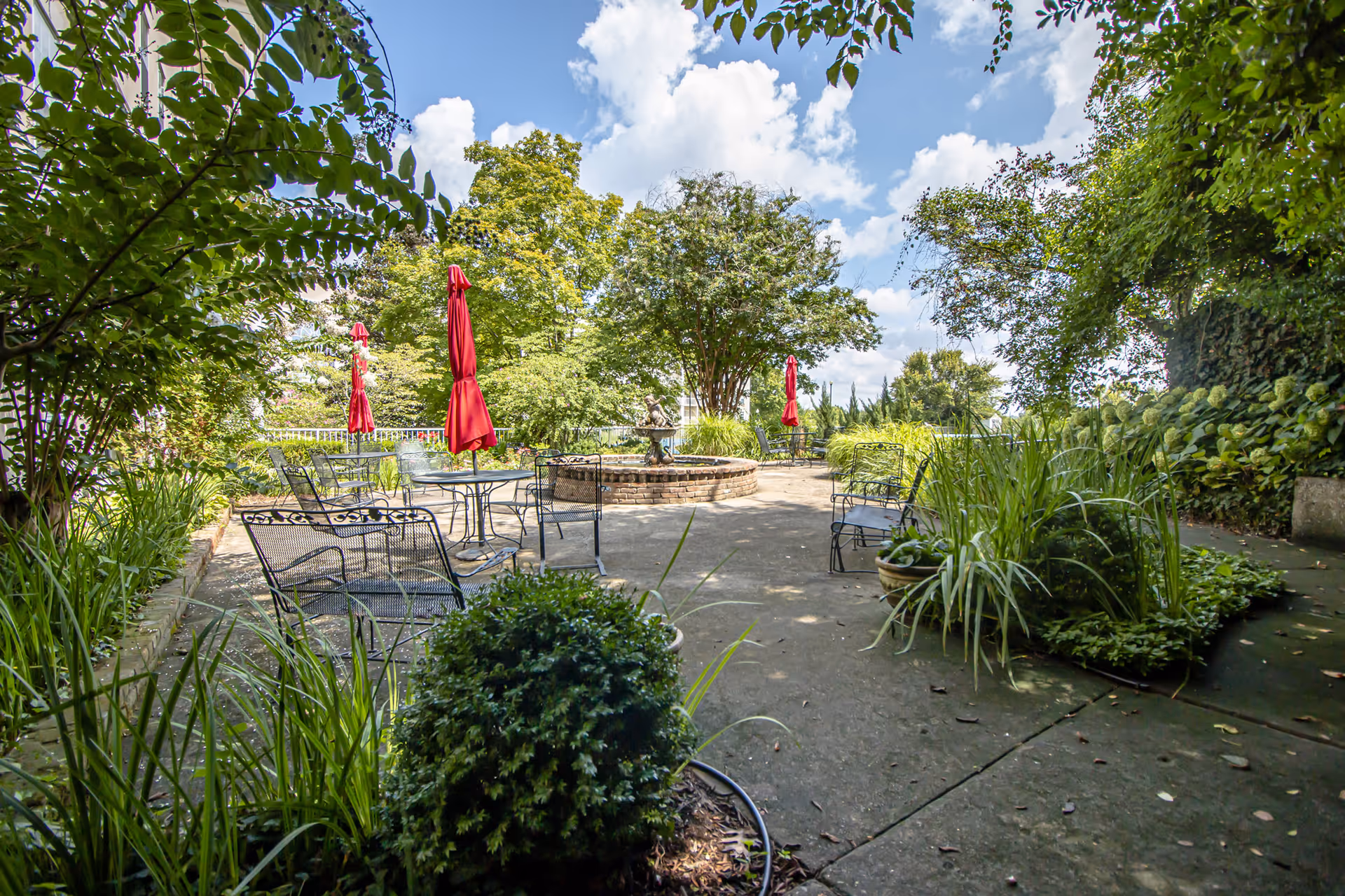 Outdoor patio area with metal chairs and tables, some with closed red umbrellas, surrounded by lush green plants and trees under a partly cloudy sky.