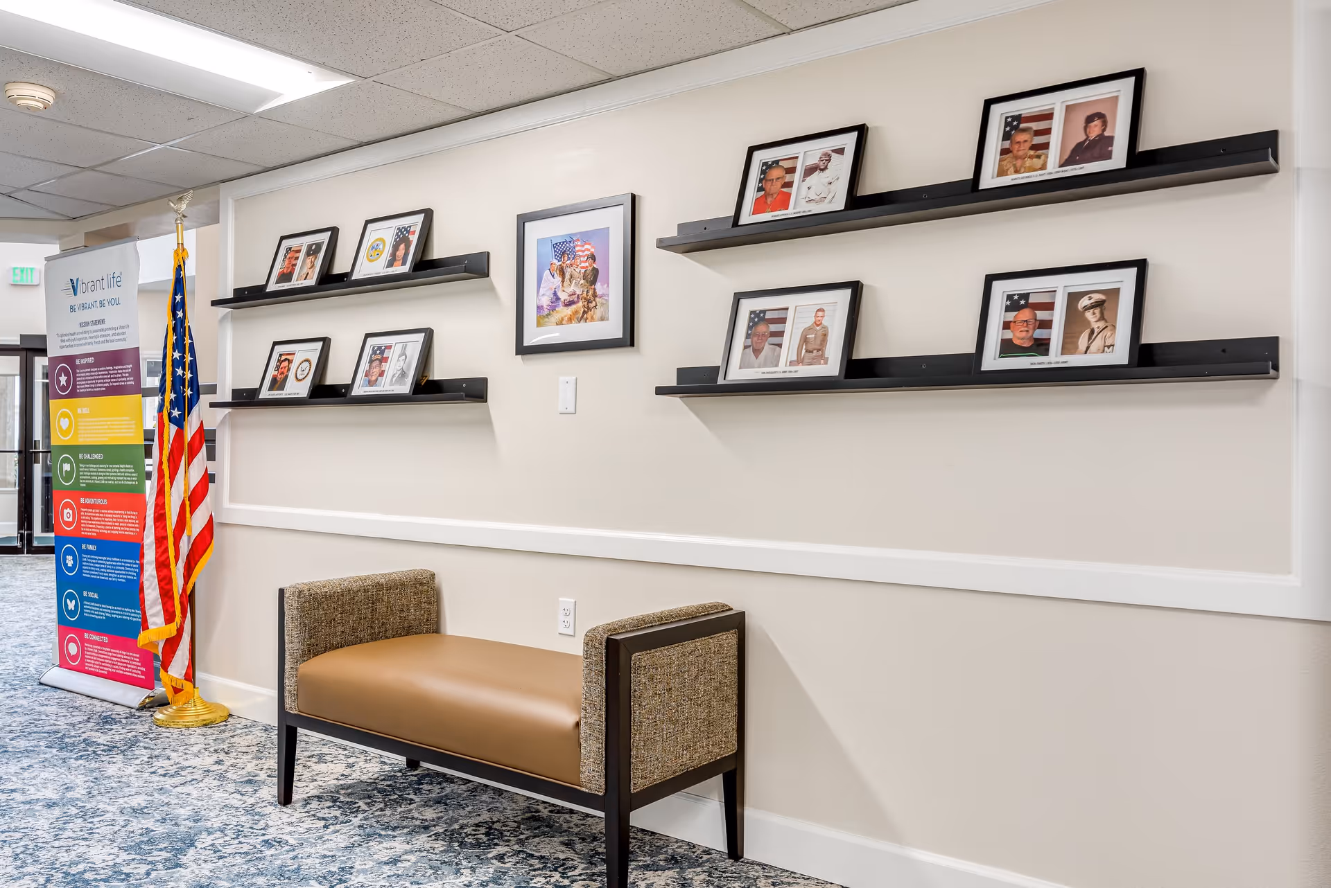 A hallway in a senior living facility with a bench against the wall. Above the bench are two black shelves displaying framed photographs, mostly portraits of veterans with American flags in the background. To the left, there is an American flag on a stand and a colorful informational banner with the title 'Vibrant Life'. The floor is carpeted with a blue and gray pattern.
