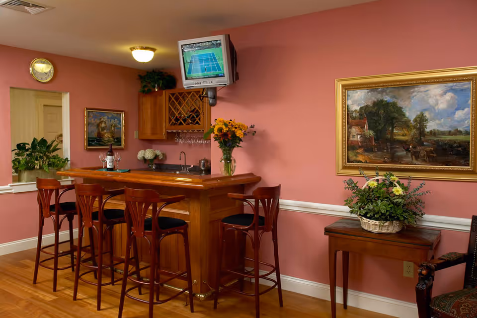 Interior view of a cozy bar area with a wooden bar counter and four high wooden chairs with black cushions. The walls are painted pink and decorated with framed paintings and a clock. A small TV is mounted on the wall above the bar, showing a tennis match. There are plants and flowers placed on the bar and a small wooden table nearby.