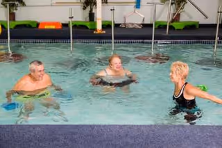 Three seniors enjoying water exercise in an indoor swimming pool, with one instructor guiding two participants in the water.
