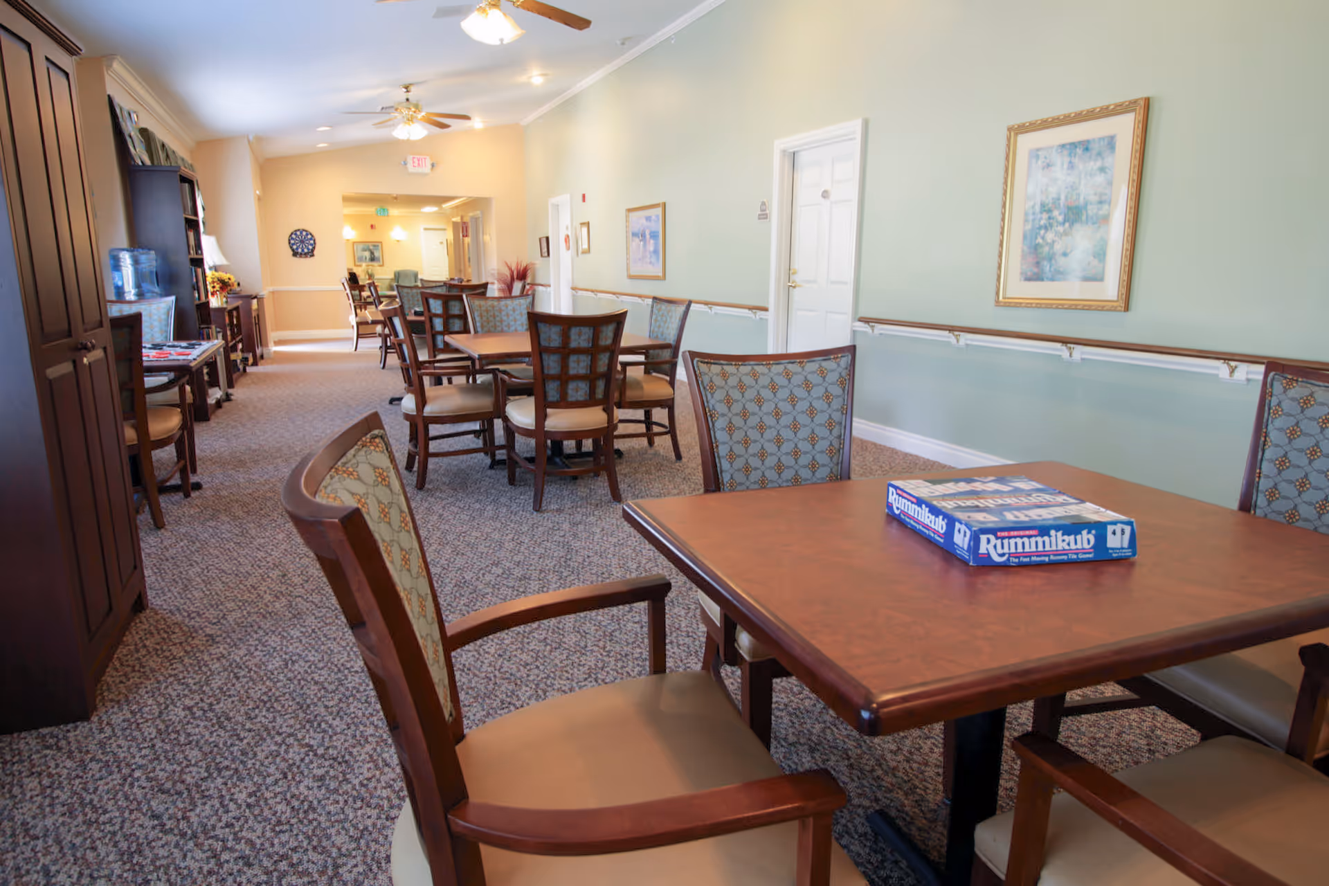 A well-lit common area in a senior living facility with multiple wooden tables and cushioned chairs arranged along a carpeted hallway. A Rummikub board game is placed on the nearest table. The walls are painted light green and beige, decorated with framed artwork. Ceiling fans and lights are visible overhead, and there is a water dispenser and bookshelf along the left wall.