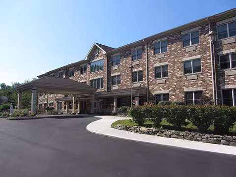 Exterior view of a three-story brick building with multiple windows and a covered entrance driveway, surrounded by landscaped bushes and a clear blue sky.