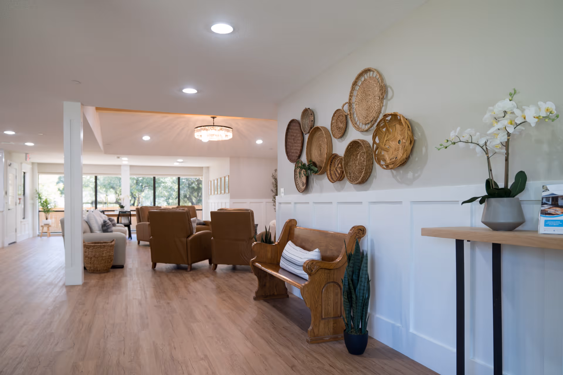 A bright and spacious living area in an assisted living facility featuring wooden flooring, a wooden bench with a striped cushion, a potted plant, and a wall decorated with various woven baskets. In the background, there are comfortable armchairs and a sofa near large windows letting in natural light, with a chandelier hanging from the ceiling.