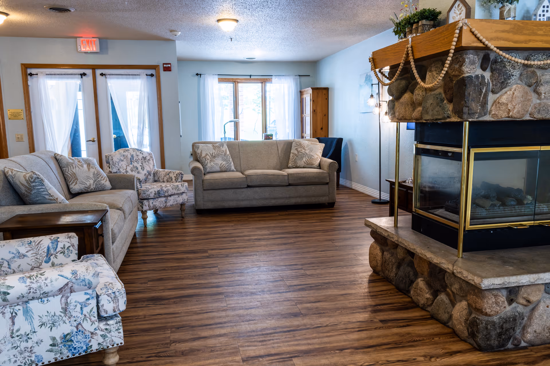 A cozy living room in a senior living facility featuring two beige sofas with patterned cushions, two floral armchairs, a stone fireplace with a glass front and wooden mantle decorated with plants and ornaments, wooden flooring, and large windows with sheer white curtains allowing natural light to fill the room.