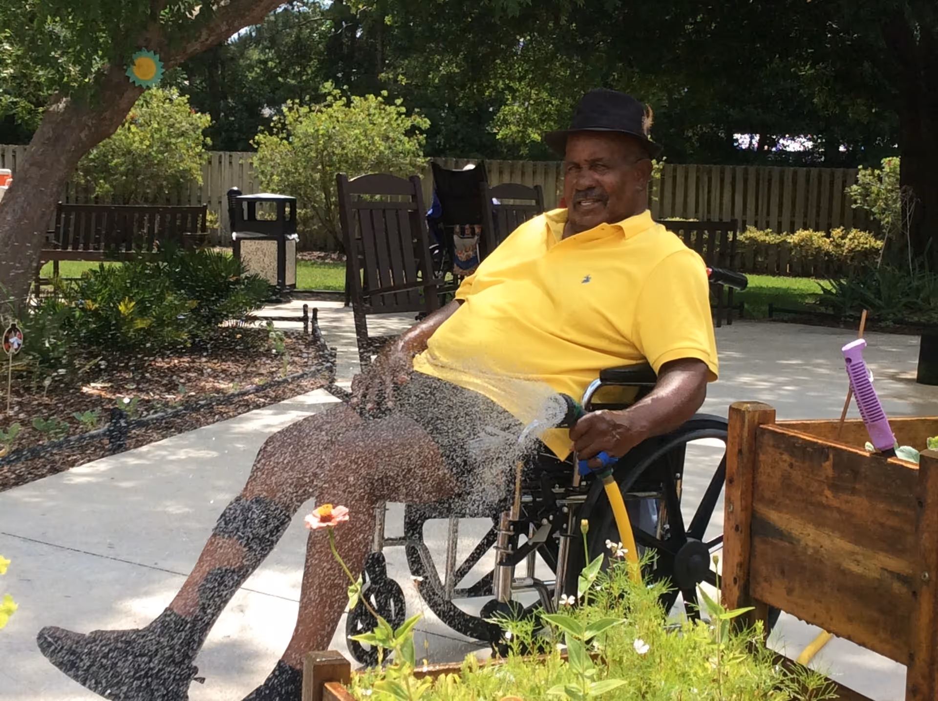 An elderly man in a wheelchair wearing a yellow shirt and black hat waters plants with a garden hose in an outdoor garden area with trees, benches, and a wooden fence in the background.