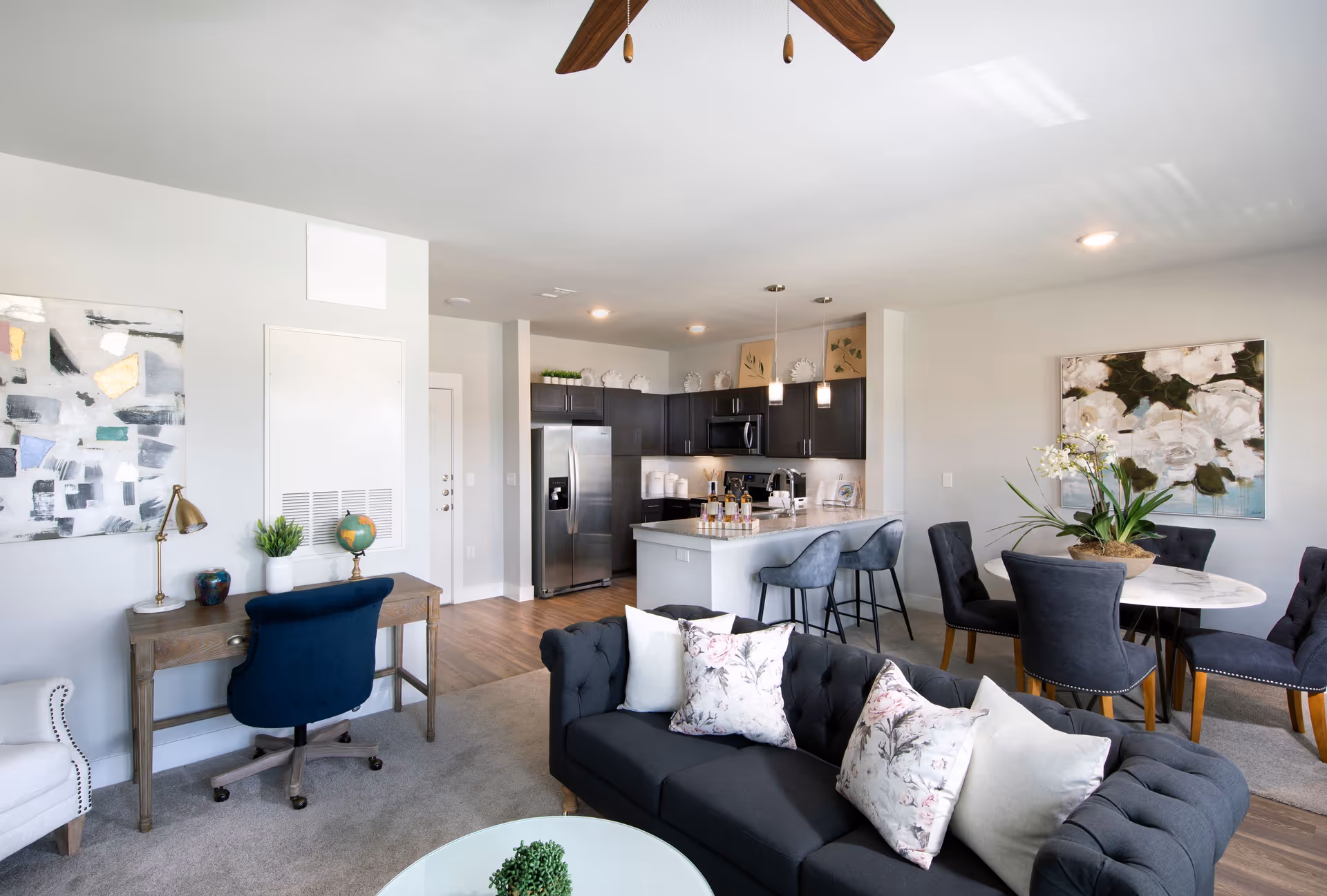 Open-concept living area showing a dark tufted sofa with floral pillows, a kitchen island with barstools, dining table, and a small desk.