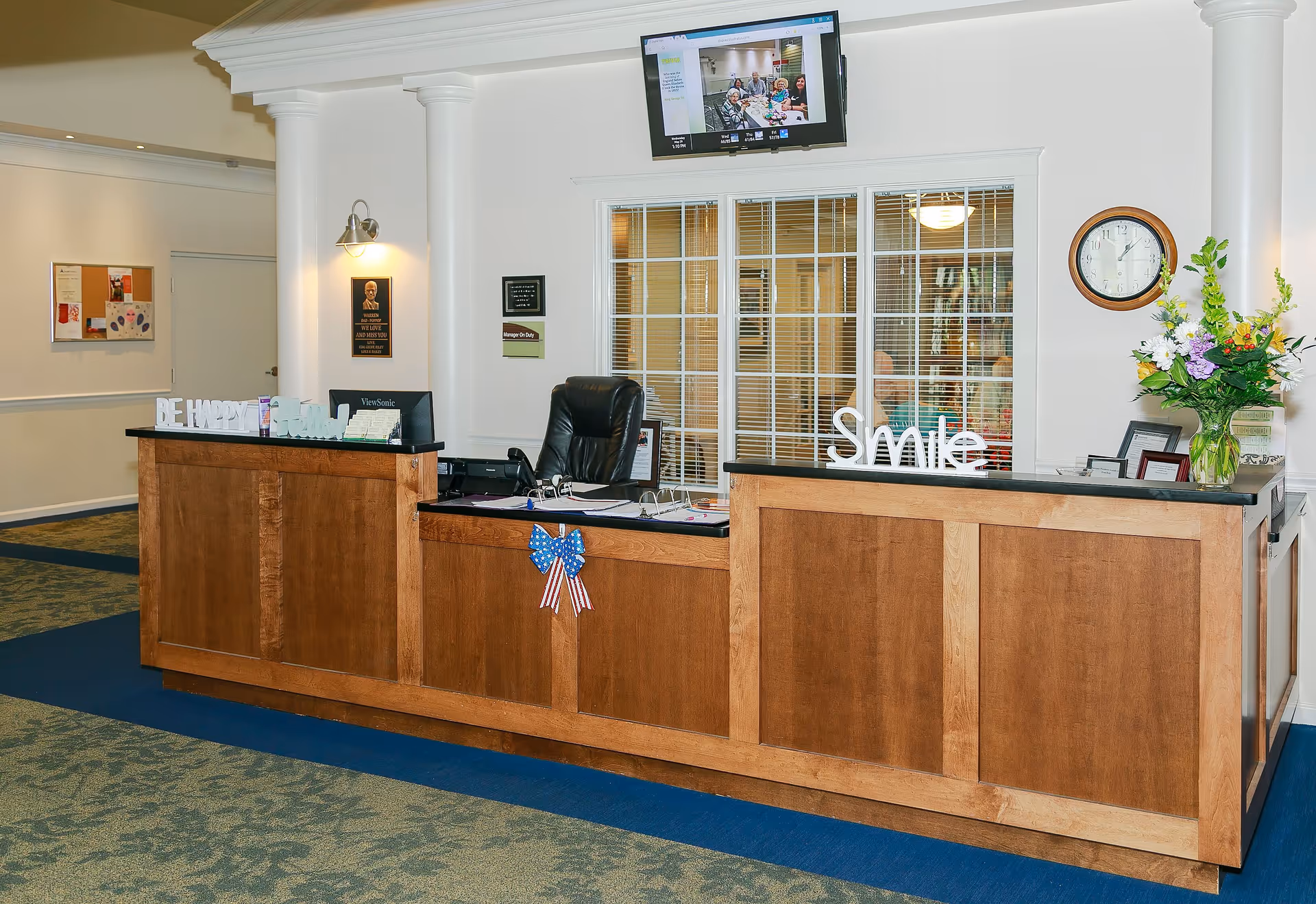 Reception desk area in a senior living facility with wooden counters, a black office chair, a computer monitor, and decorative items including a 'Smile' sign and a patriotic bow. Behind the desk is a window with blinds, a wall clock, a vase of flowers, and a TV mounted on the wall displaying a video call with several people.