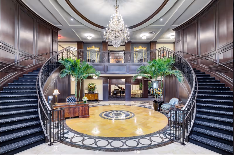 Elegant interior lobby with a grand double staircase curving upwards on both sides, dark wood paneling on the walls, a large chandelier hanging from the ceiling, two tall potted plants flanking a wooden reception desk, and a circular patterned floor design in the center. In the background, a grand piano and seating area are visible.