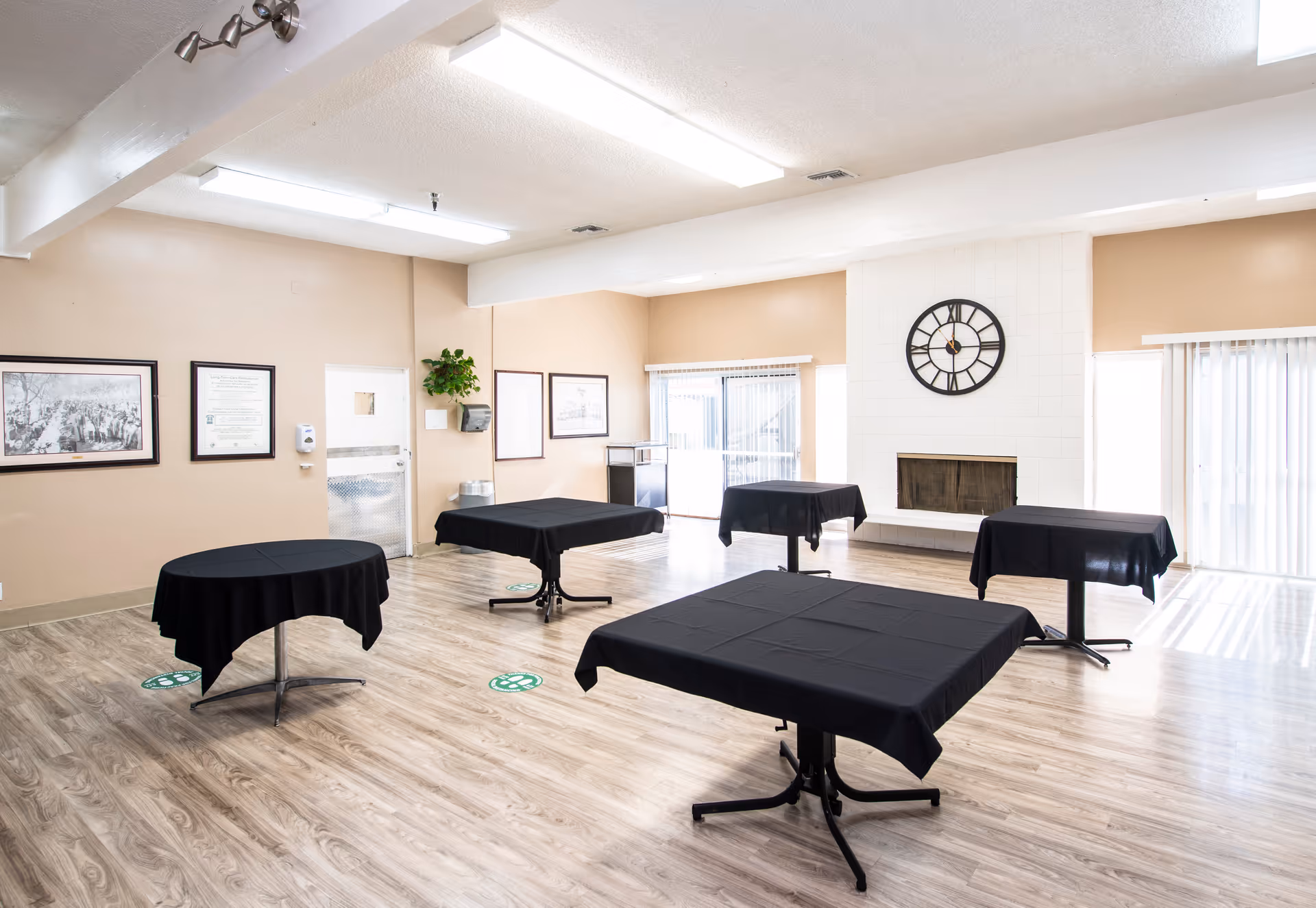 A spacious room with light wood flooring and beige walls featuring five tables covered with black tablecloths spaced apart. The room has a white tiled fireplace with a large black clock above it, framed pictures on the walls, and large windows with vertical blinds allowing natural light to enter.