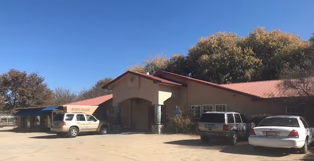 Single-story building with a red metal roof and front entrance, a small awning reading 'green house', and several parked cars under a clear blue sky.