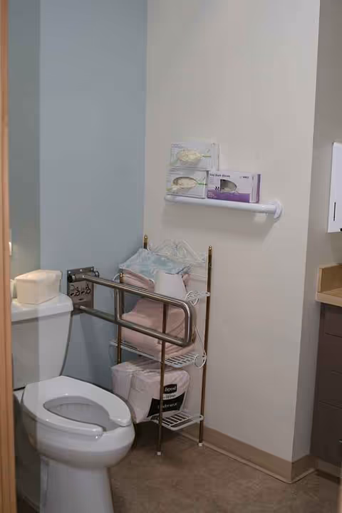 Bathroom in a senior living facility with a toilet, safety rails, towels, and shelf units.