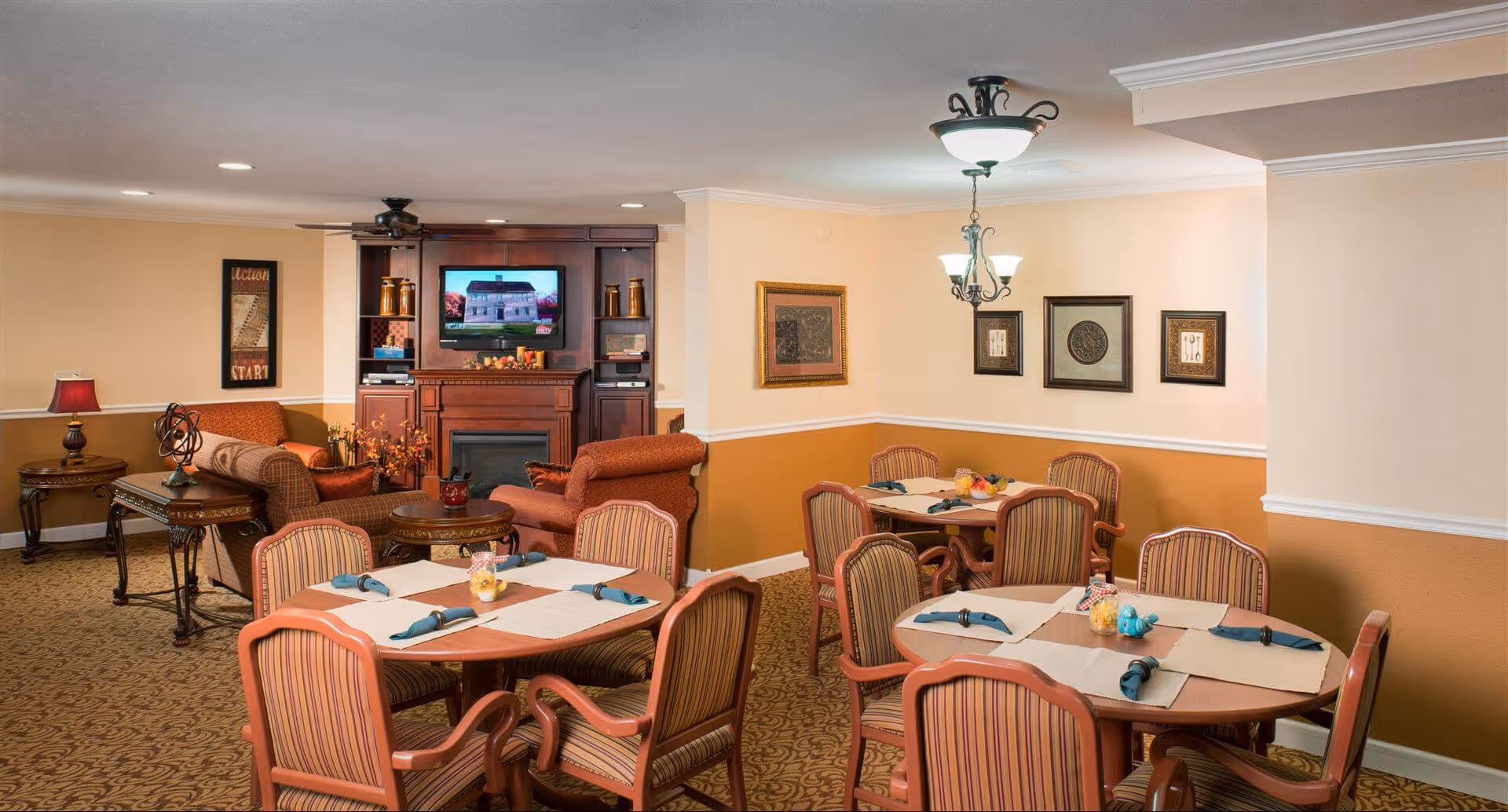 Communal dining and lounge area with round tables set for meals, upholstered chairs, and a TV above a fireplace.