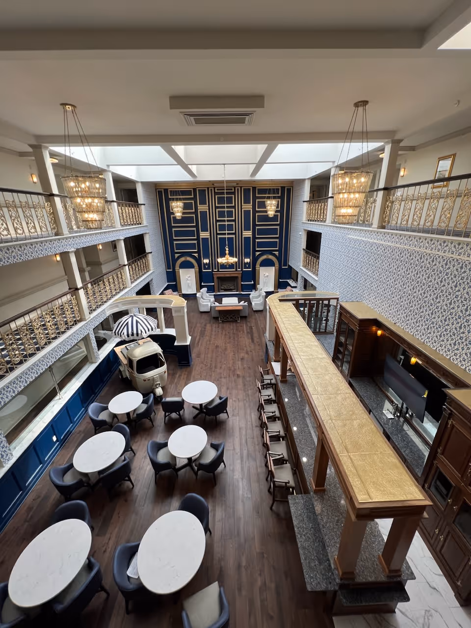 Spacious and elegant common area with multiple round tables and chairs arranged on a wooden floor. The room features a high ceiling with large chandeliers, ornate railings on the upper balconies, and a decorative blue and gold accent wall with a fireplace and white statues. There is also a long bar counter with stools on one side and a small vintage-style vehicle with a striped umbrella near the tables.