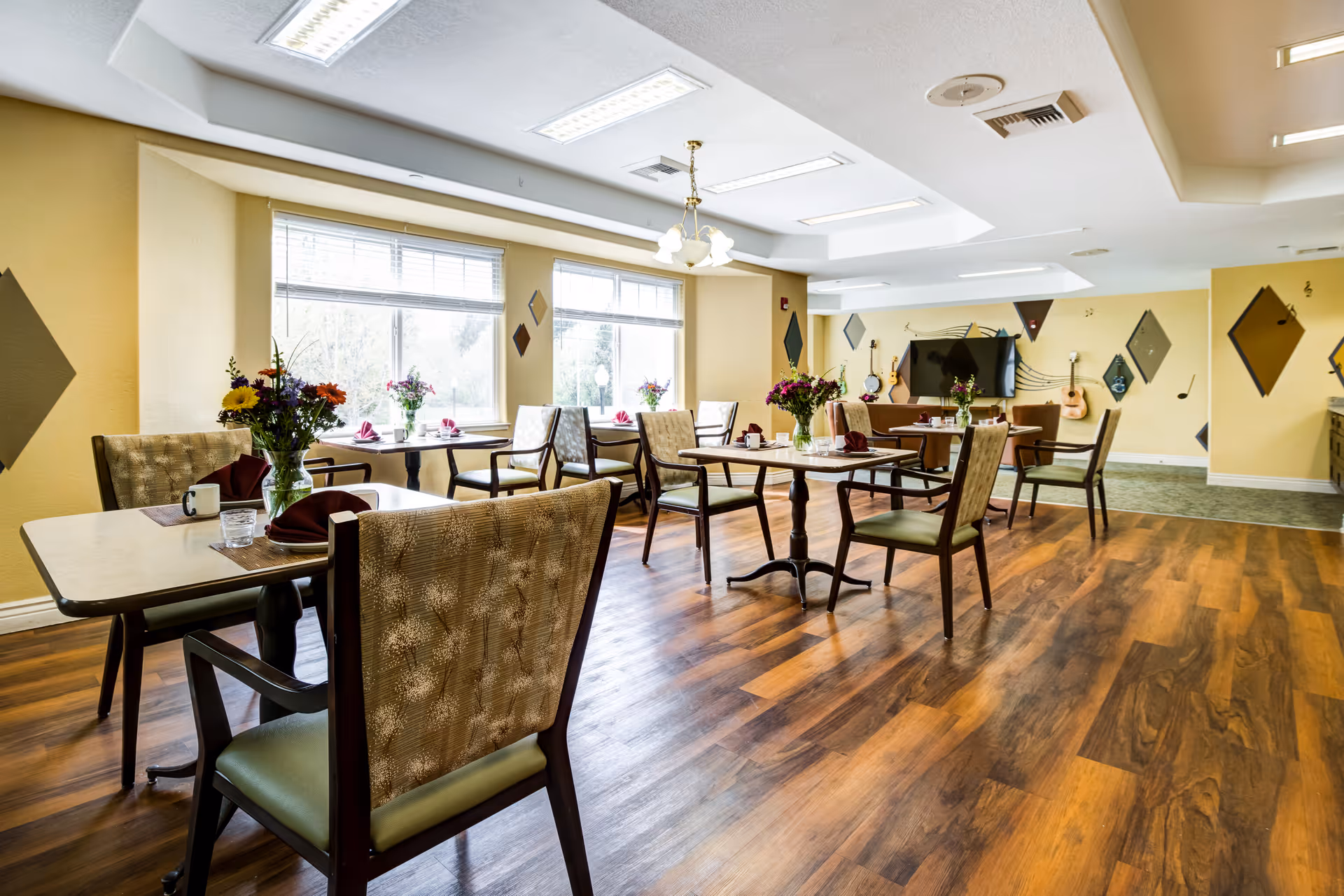 A bright dining room with several tables and chairs arranged neatly. Each table has a vase with fresh flowers and place settings with napkins and cups. Large windows let in natural light, and the walls are decorated with diamond-shaped wall art and musical notes. A television and guitars are visible in the background.