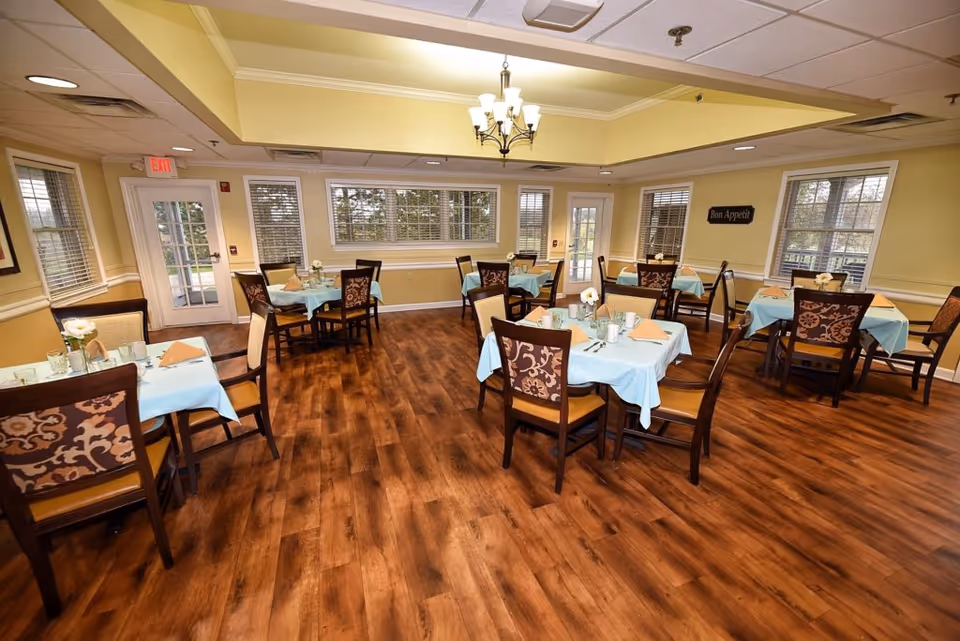 Bright dining room with several tables set with light blue tablecloths and wooden chairs on a polished wood floor.