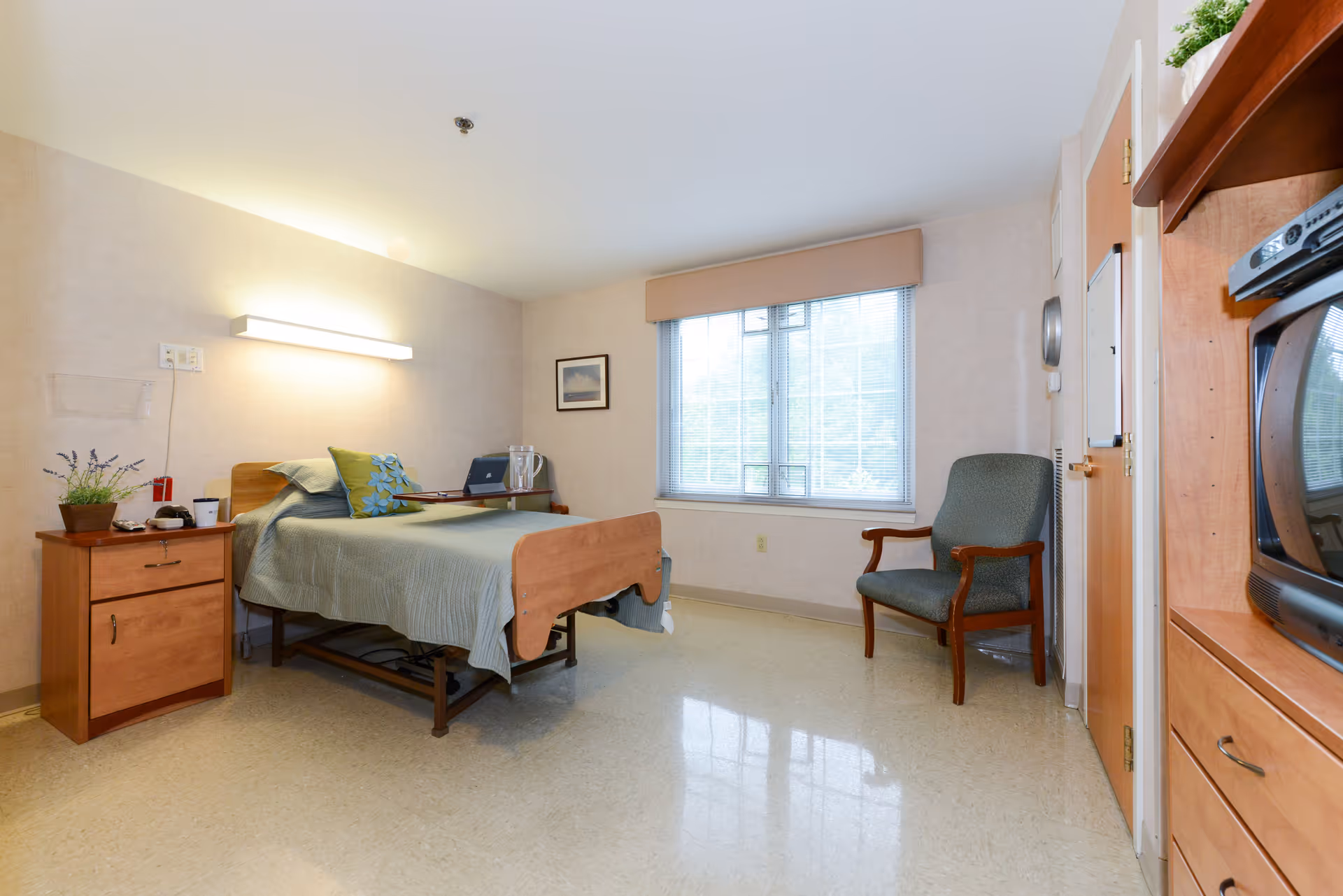 A simple, clean senior living bedroom with a single bed covered in a green quilt and decorative pillows. Next to the bed is a wooden nightstand with a plant, phone, and other small items. A large window with blinds lets in natural light. There is a comfortable armchair near the door and a wooden cabinet with a TV on the right side of the room. The walls are light-colored, and the floor is a polished beige tile.