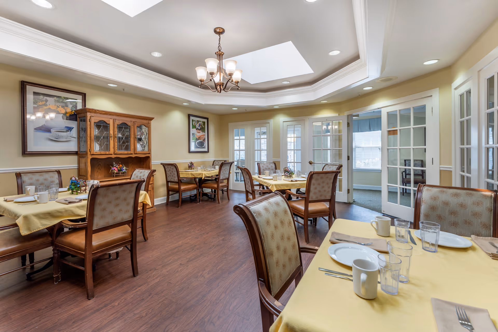 A dining room with several tables covered in yellow tablecloths, set with plates, cups, glasses, and utensils. The room features wooden chairs with cushioned seats and backs, a wooden cabinet against the wall, framed artwork, a chandelier hanging from a tray ceiling with a skylight, and French doors leading to another room.