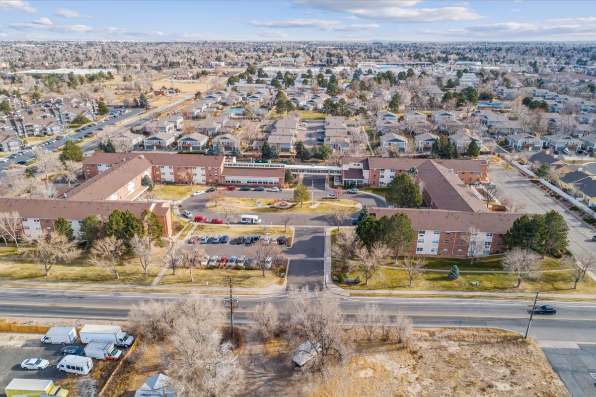 Aerial view of a large senior living facility with multiple connected brick buildings forming a U-shape, surrounded by parking lots, trees, and residential neighborhoods under a partly cloudy sky.