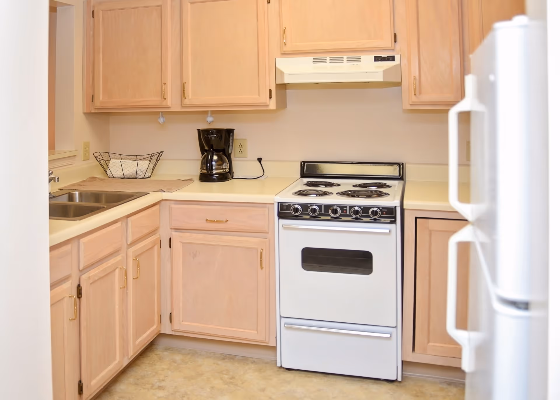 A small kitchen with light wood cabinets, a white electric stove with four burners, a white refrigerator, a double sink, a black coffee maker, and a wire basket on the countertop.