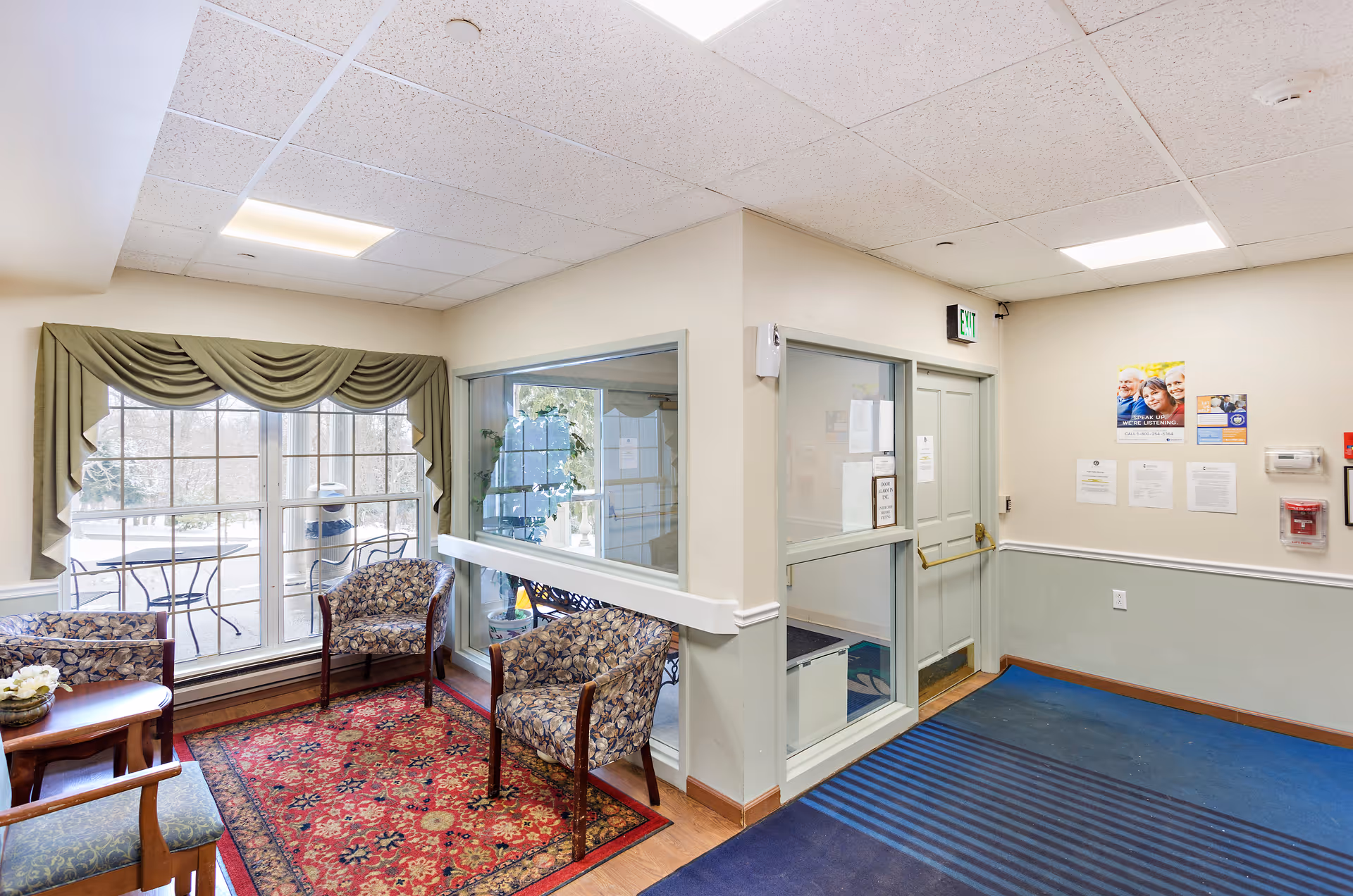 Bright senior living facility lobby with chairs, a patterned rug, large windows and an exit door.