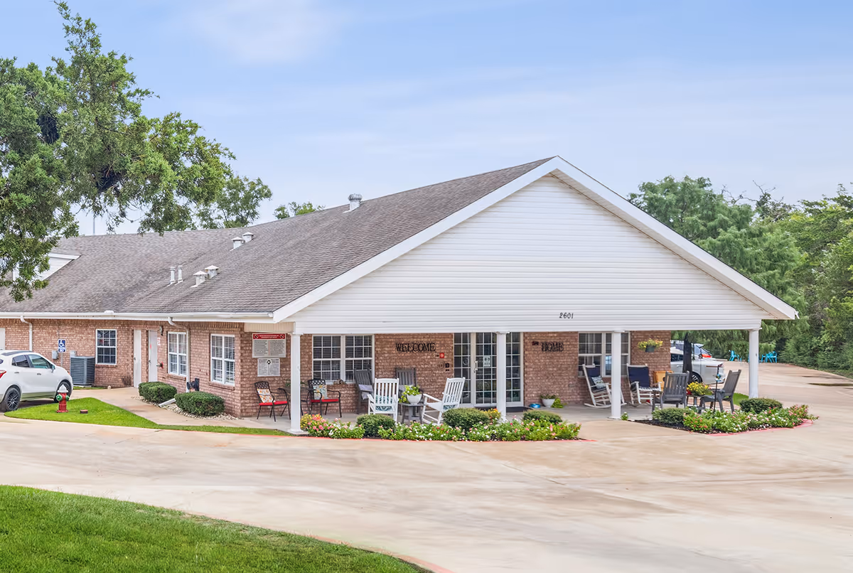 Exterior view of a single-story brick building with a large white covered porch area featuring several chairs and small tables. The building is surrounded by a paved driveway and landscaped with green grass, bushes, and flowers. Trees are visible in the background under a clear sky.