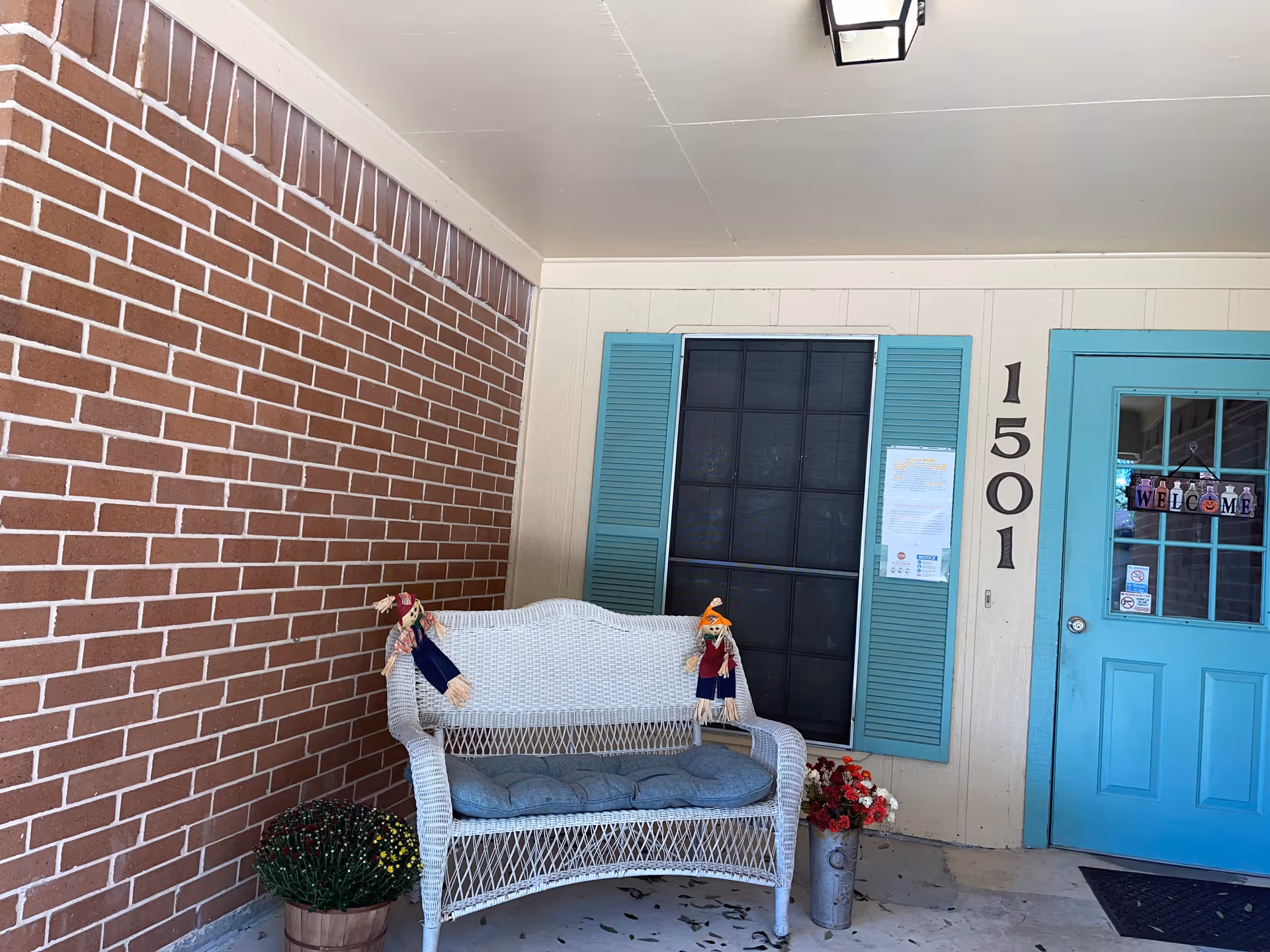 Front porch entrance with a turquoise door numbered 1501, a white wicker bench, shuttered window, and potted plants.