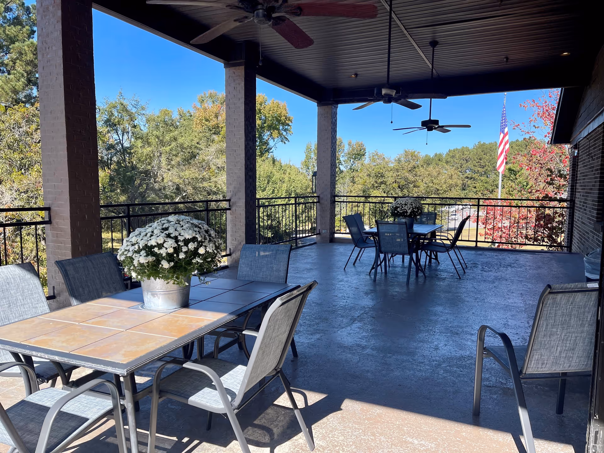 Covered outdoor patio area with two tables, each surrounded by chairs. Each table has a pot of white flowers. Ceiling fans hang from the roof. Trees and a clear blue sky are visible in the background along with an American flag on a flagpole.