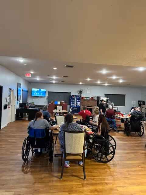 A group of elderly individuals, some in wheelchairs, seated around tables in a spacious room with wooden floors and white walls. A television is mounted on the wall, and a staff member is standing near the back of the room. The room is well-lit with ceiling lights and has a cozy, communal atmosphere.