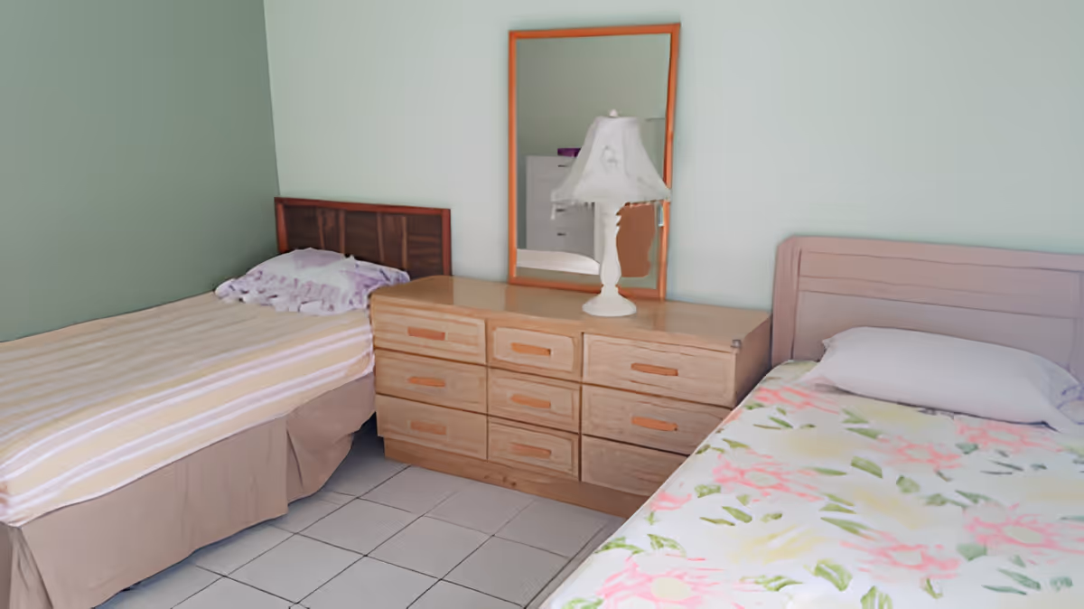 A bedroom with two single beds separated by a wooden dresser with multiple drawers. On top of the dresser is a white table lamp and a rectangular mirror hanging on the wall. The bed on the left has a striped beige and white bedspread with a purple pillow, while the bed on the right has a floral bedspread with a white pillow. The walls are painted light green and the floor is tiled.