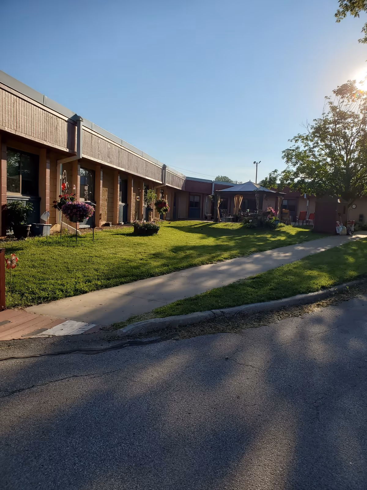Outdoor view of a senior living facility with a single-story building, a well-maintained lawn, hanging flower baskets, a paved walkway, and a gazebo with outdoor seating under a clear blue sky during daylight.