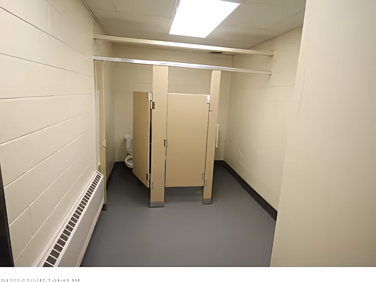 A small public restroom with beige walls and a gray floor. There are two beige stall doors, one partially open showing a white toilet inside. A white radiator is mounted on the left wall, and a rectangular overhead light fixture illuminates the room.