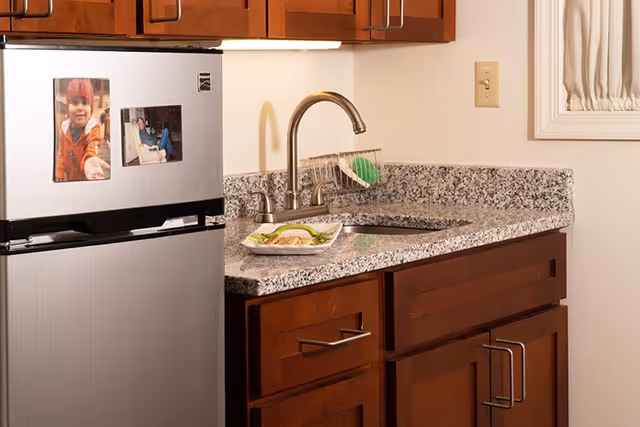 A small kitchen area with a stainless steel mini-fridge with photos, granite countertop, sink, and dark wood cabinets.