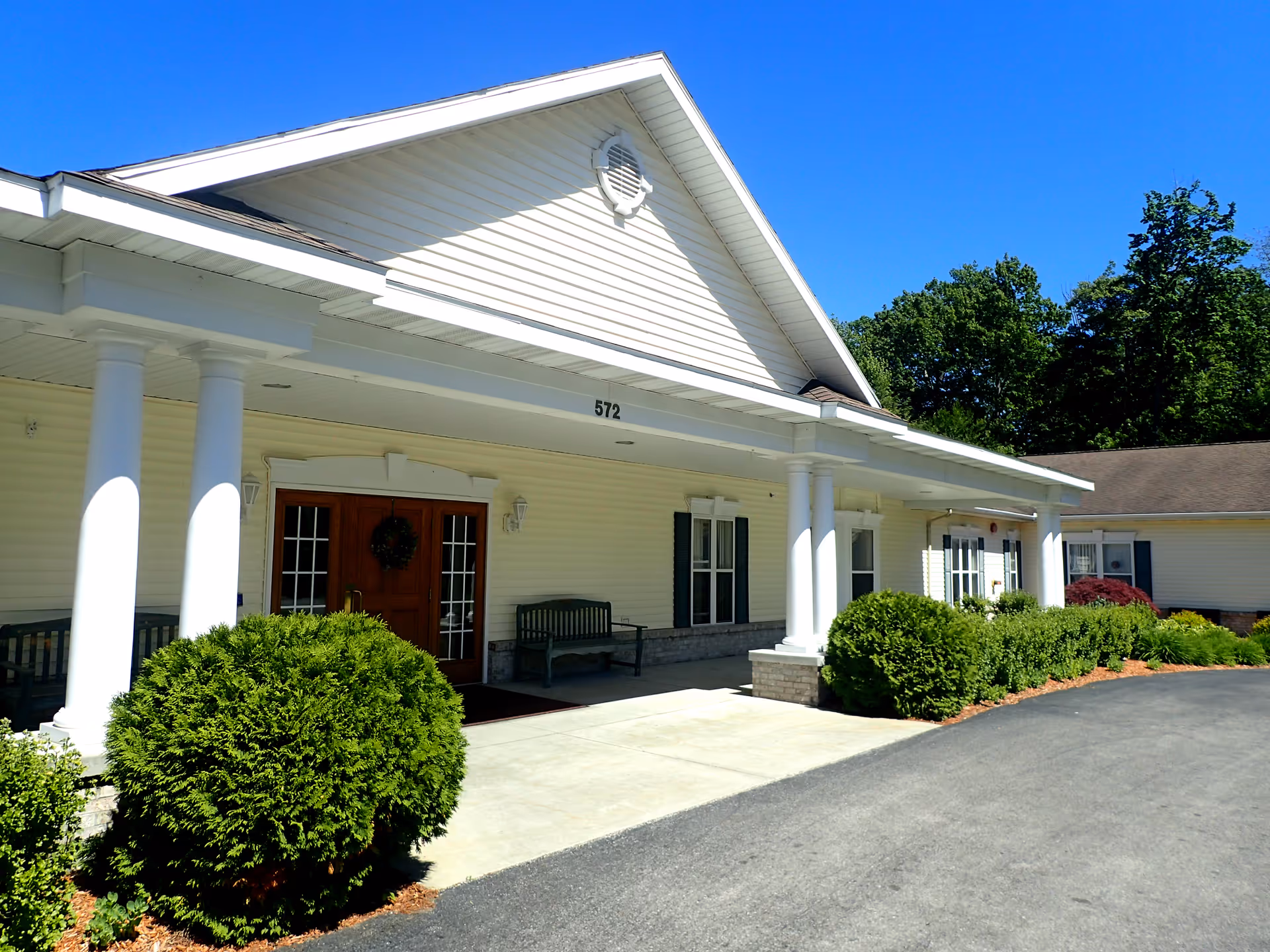 Front exterior view of a single-story assisted living facility building with white siding, a peaked roof, and a covered entrance supported by white columns. There are benches near the entrance, green bushes along the walkway, and trees in the background under a clear blue sky.