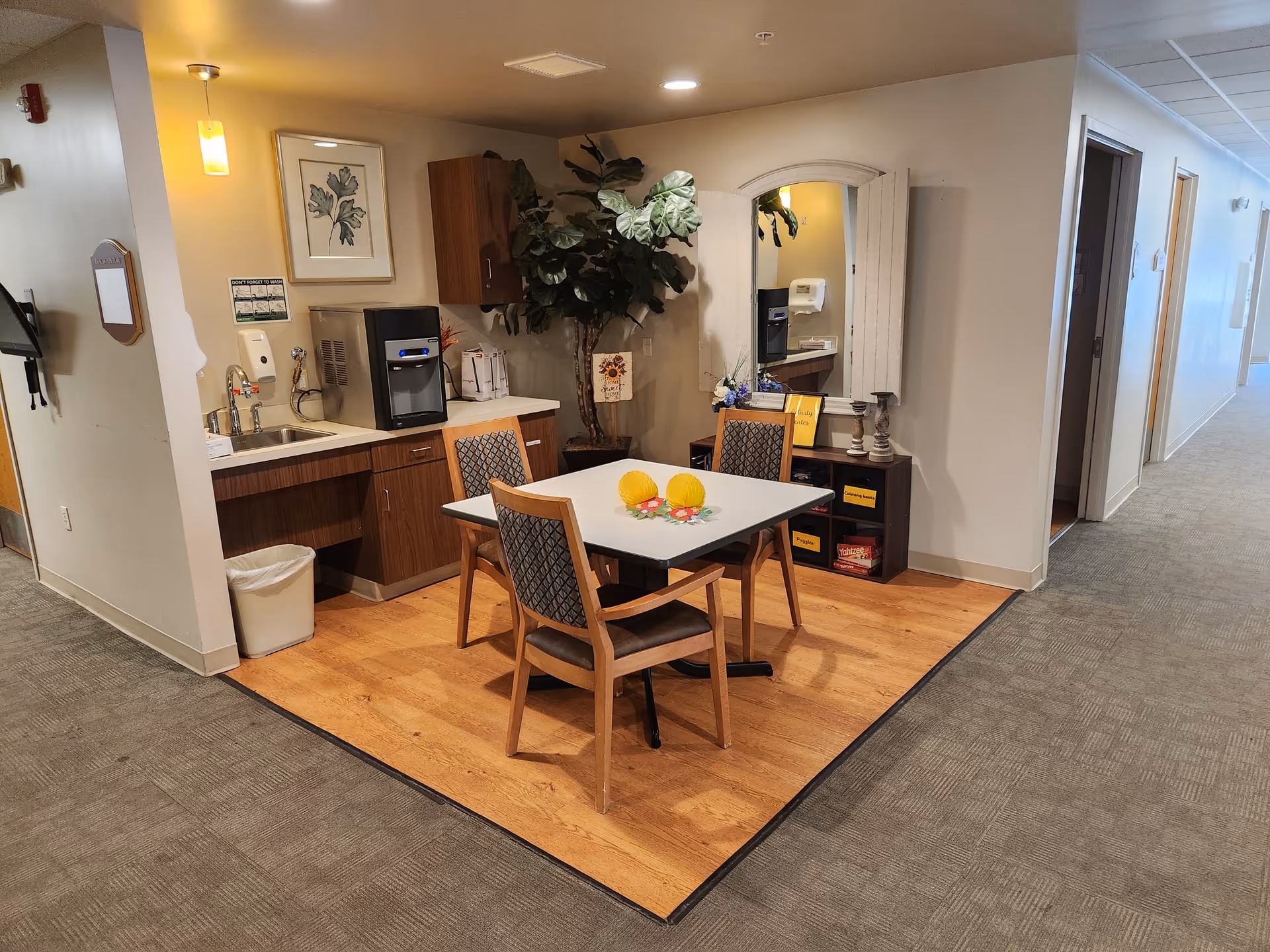 A small seating area in a senior living facility with a square table and four wooden chairs with patterned cushions. The area has a wood floor section contrasting with the surrounding carpet. There is a kitchenette with a sink, water dispenser, and cabinets. A large potted plant and a wall-mounted mirror with shutters are visible. The hallway extends to the right with several doors.