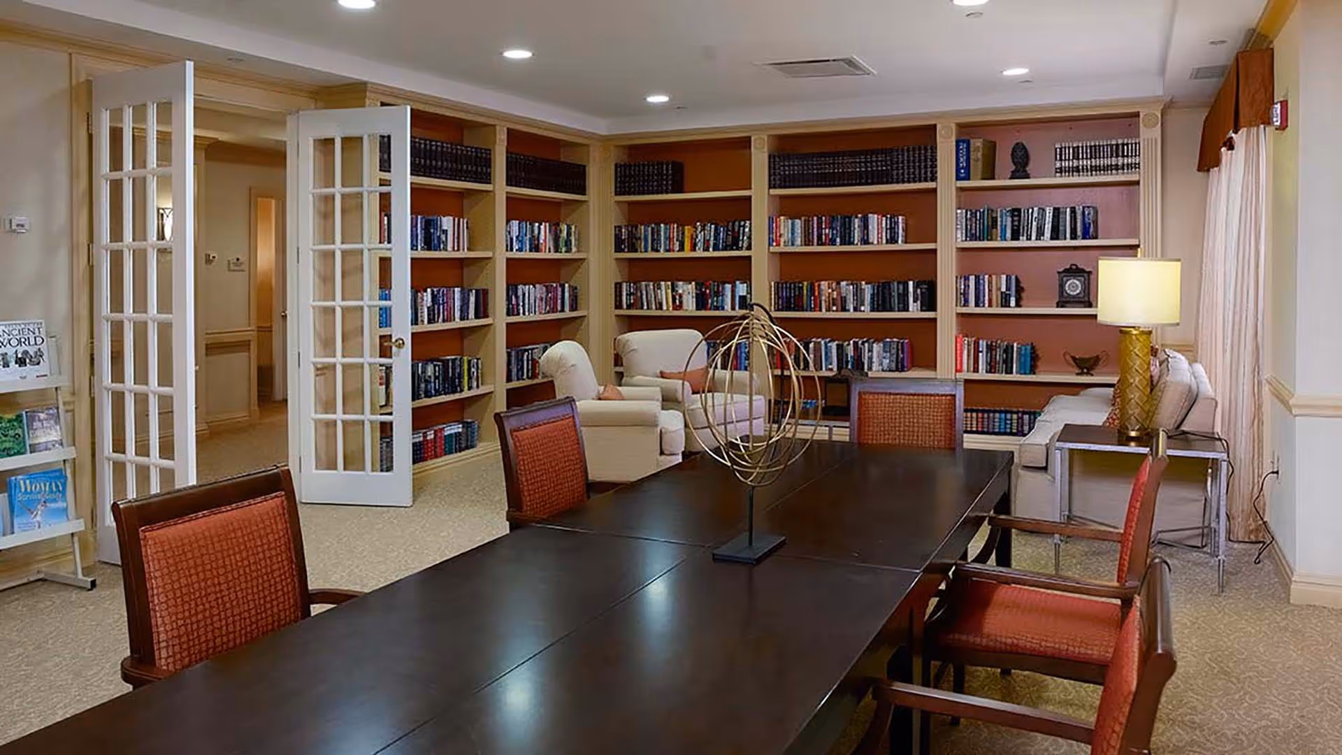 A cozy library or reading room with a large dark wooden table surrounded by red cushioned chairs. In the background, there are built-in bookshelves filled with books, two beige armchairs, a beige sofa, and a side table with a lamp. French doors lead to a hallway, and a small book display rack is visible on the left side.
