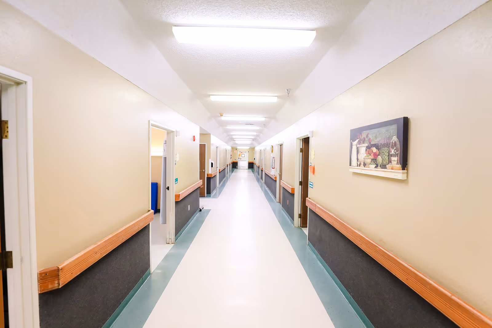 A long, brightly lit corridor of a rehabilitation facility with wall handrails, open doorways, and framed artwork.