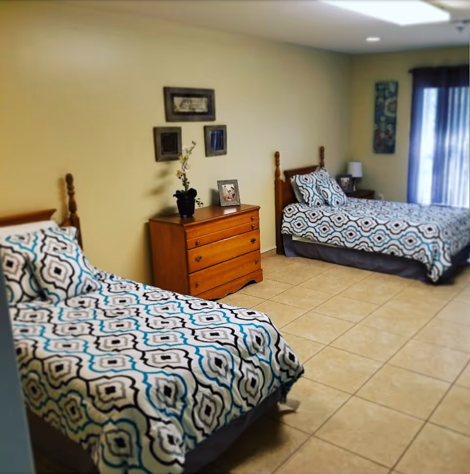 Two twin beds with blue-and-white patterned bedding, a wooden dresser, and a curtained window in a tiled bedroom.