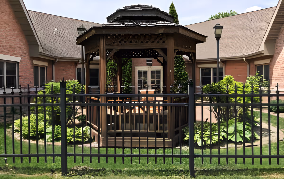 Outdoor view of a courtyard at Villas at Kewanee featuring a wooden gazebo with a table and chairs inside, surrounded by a black metal fence, greenery, bushes, and brick buildings with windows and sloped roofs in the background.