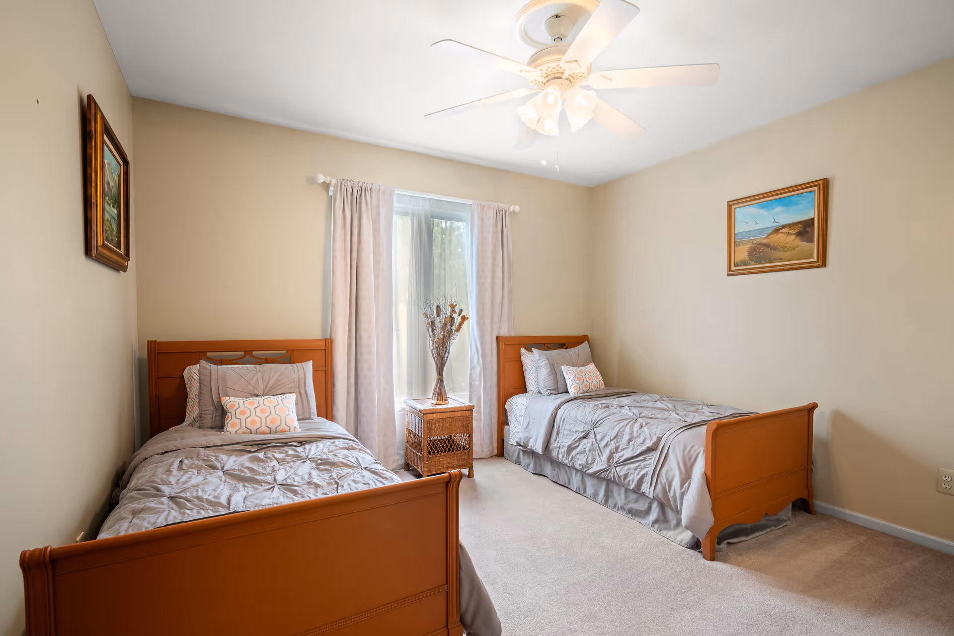 A bedroom with two single wooden beds, each made with gray bedding and decorative pillows. Between the beds is a wicker nightstand with a vase of dried flowers. The room has beige walls, a window with light curtains, a ceiling fan with lights, and two framed paintings on the walls.