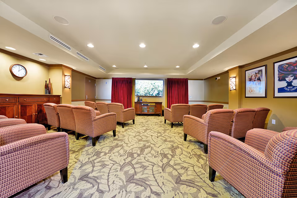 A cozy media room with multiple upholstered armchairs arranged in rows facing a large flat-screen TV mounted on the wall. The room has patterned carpet, warm yellow walls, framed artwork, a wooden cabinet, and recessed ceiling lights.