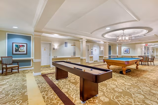Interior view of a recreational room in a senior living facility featuring a shuffleboard table and a pool table on a patterned carpet. The room has beige walls with white trim, decorative columns, framed artwork, and several chairs along the walls. Ceiling lights and a chandelier provide illumination.