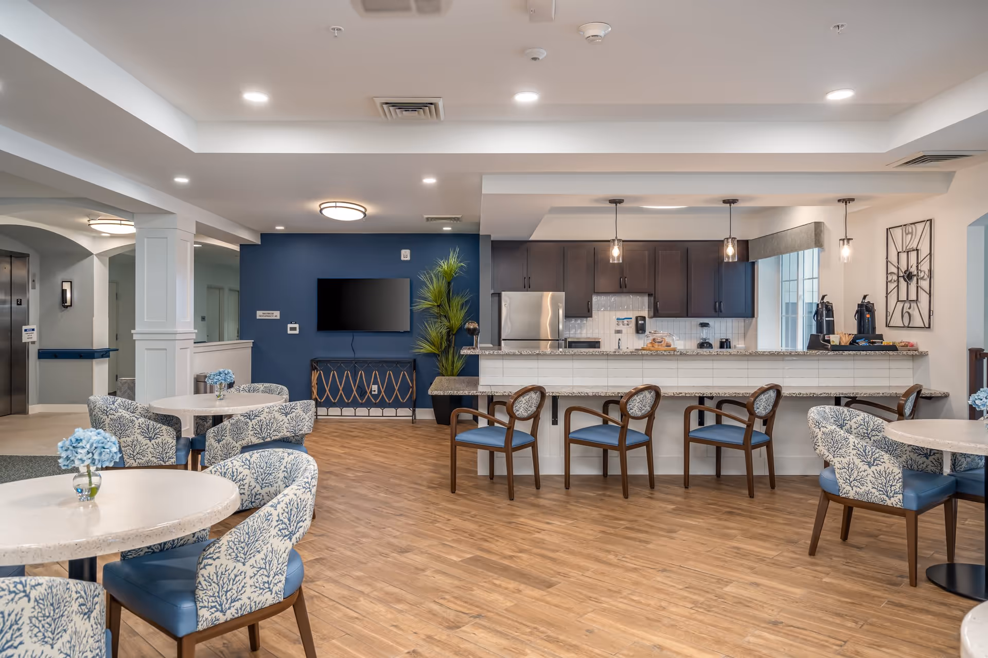 A modern senior living community common area featuring a kitchen with dark wood cabinets, stainless steel refrigerator, and a granite countertop bar with four wooden chairs. The room has light wood flooring, round tables with blue and white patterned chairs, blue accent wall with a mounted flat-screen TV, and decorative plants. Pendant lights hang over the kitchen counter, and a large decorative wall clock is visible on the right.