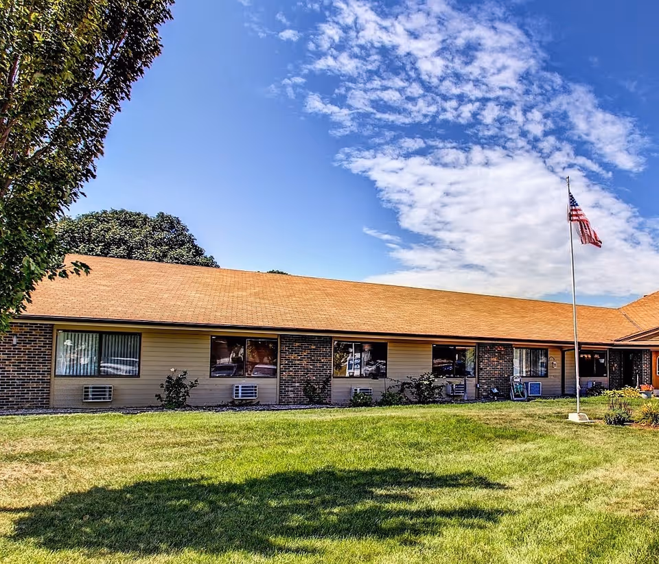 Single-story brick senior living building with windows, a front lawn and an American flag on a flagpole under a partly cloudy sky.
