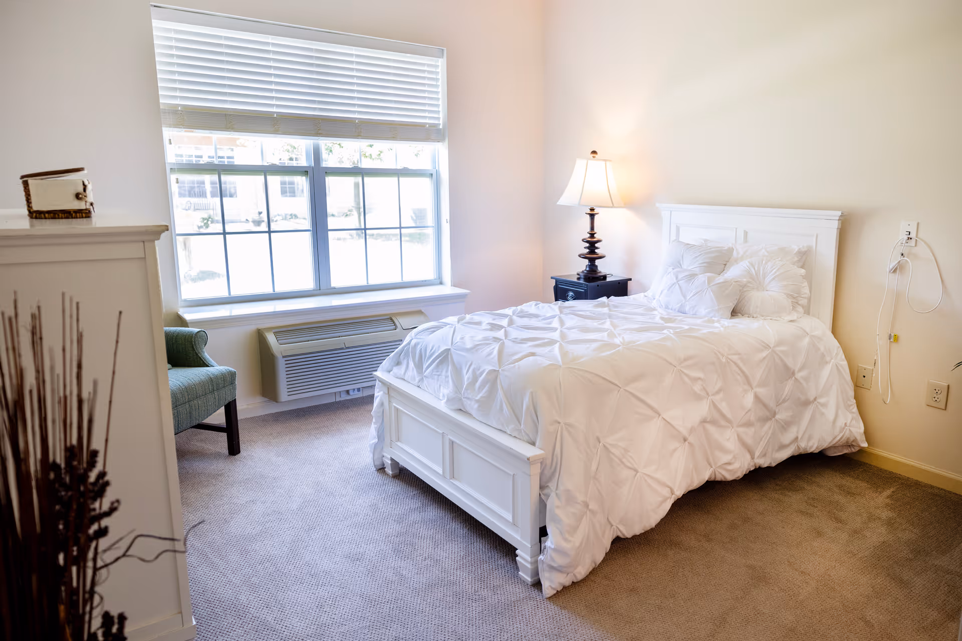 A bright bedroom with a white bed frame and white bedding, a black nightstand with a lamp, a large window with blinds, a green upholstered chair, and a white dresser with decorative items on top.