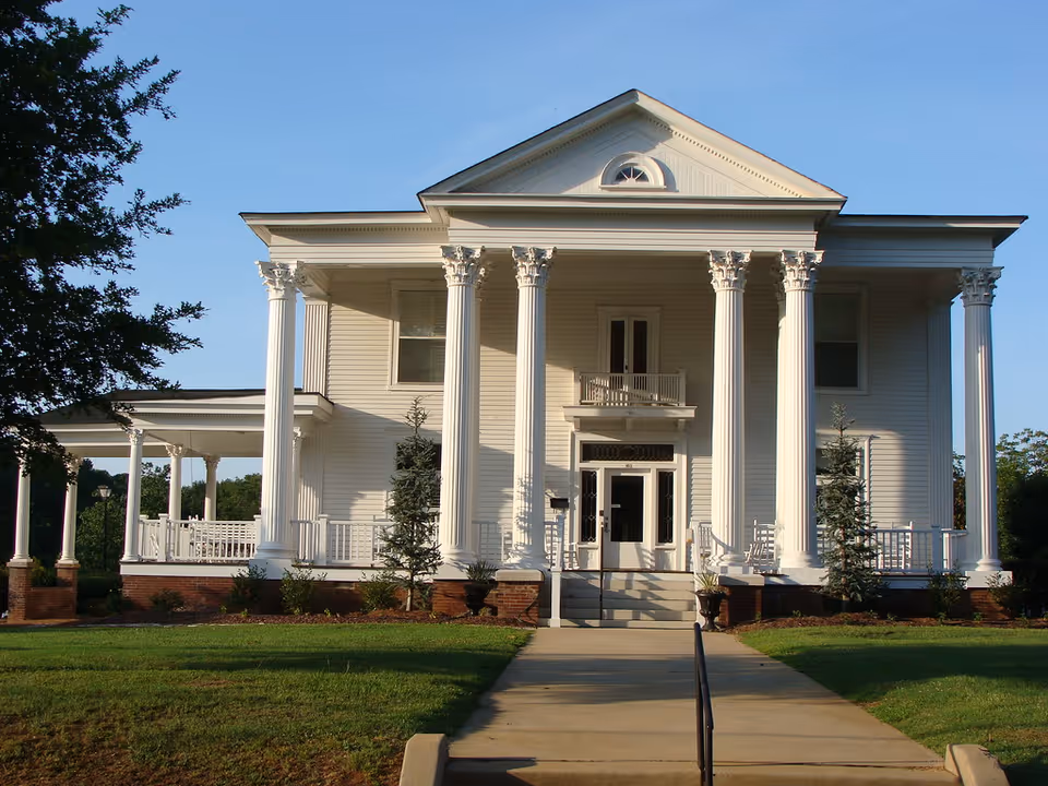 Front exterior view of a large white two-story building with tall classical columns and a porch, surrounded by a lawn and trees under a clear blue sky.