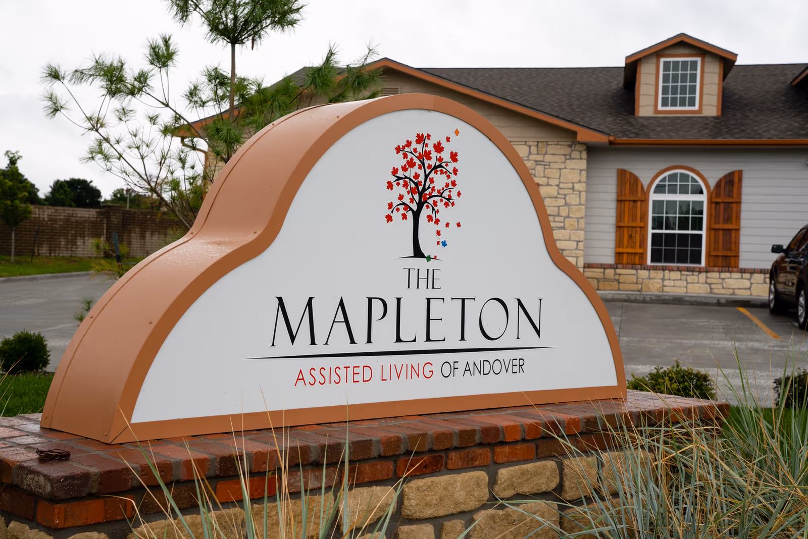 A large sign for The Mapleton Assisted Living of Andover with a tree logo featuring red and a few colored leaves, set on a brick and stone base in front of a building with stone and wood exterior and a parking lot.