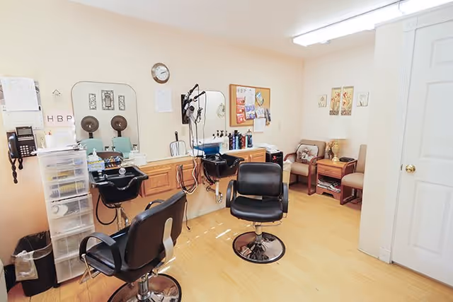 Interior of a hair salon area in a senior living facility with two black salon chairs in front of two black hair washing sinks. There are mirrors, hair care products, and a wall-mounted hair dryer. The room has light-colored walls and wooden flooring. In the corner, there are two cushioned chairs with a small table between them, decorated with a lamp and framed pictures on the wall.