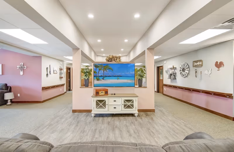 Bright communal living area with a TV displaying a beach scene on a white console, seating and decorative wall accents.