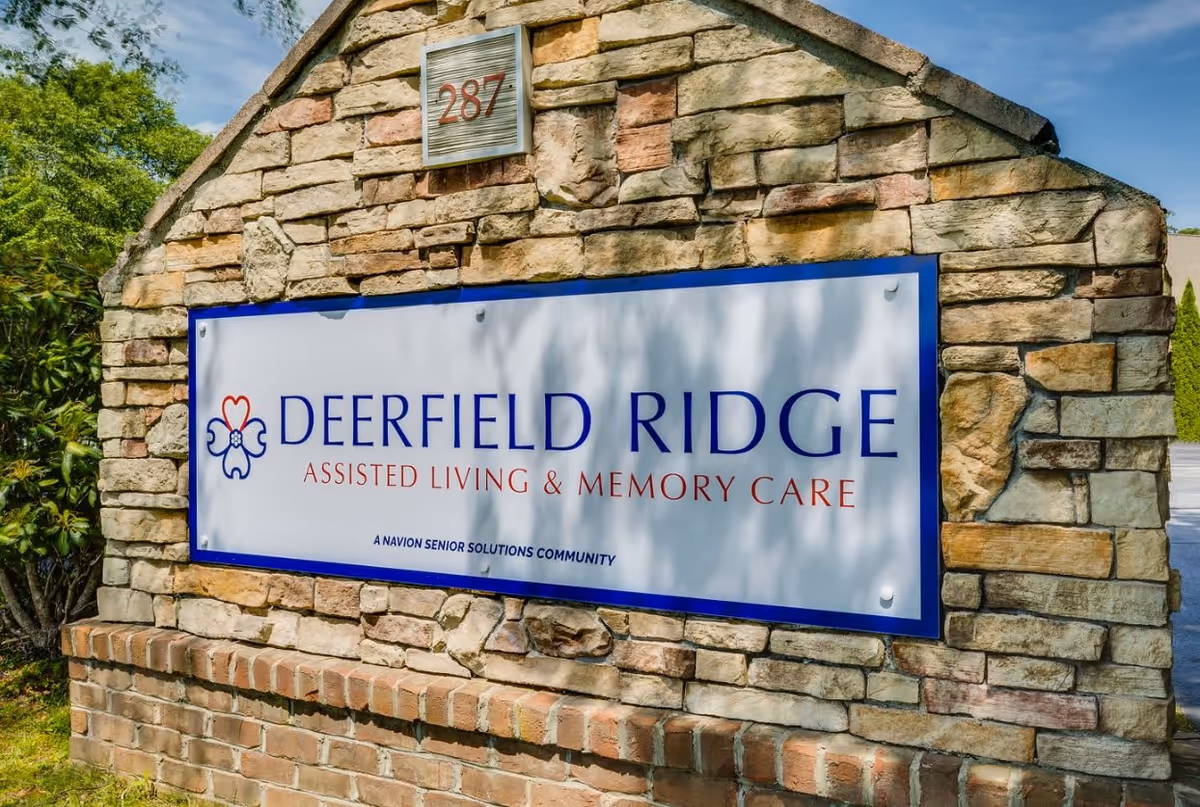 Stone sign for Deerfield Ridge Assisted Living & Memory Care with the address number 287 on top, surrounded by greenery and a clear blue sky.