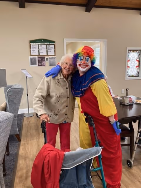 An elderly woman with white hair and glasses, wearing a beige jacket and red pants, stands next to a person dressed as a colorful clown with a rainbow wig, red nose, and face paint. Both are smiling and posing together inside a senior living facility common area with chairs, tables, and a bulletin board in the background.